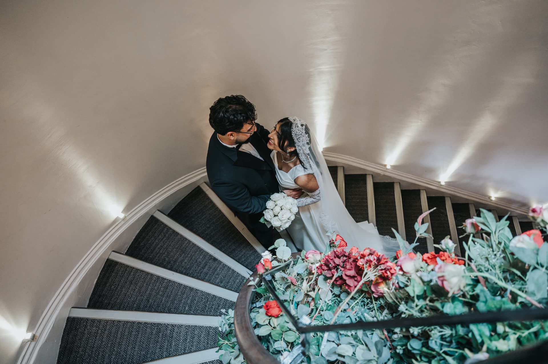 Bride and groom embracing on a curved staircase beneath floral arrangements