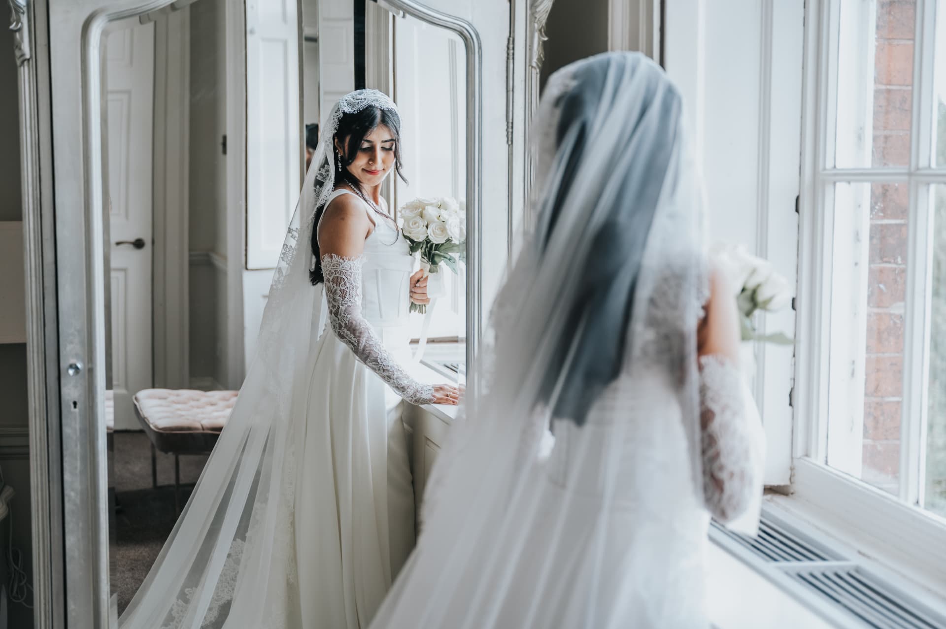 Bride holding white roses beside a full-length mirror and bright window