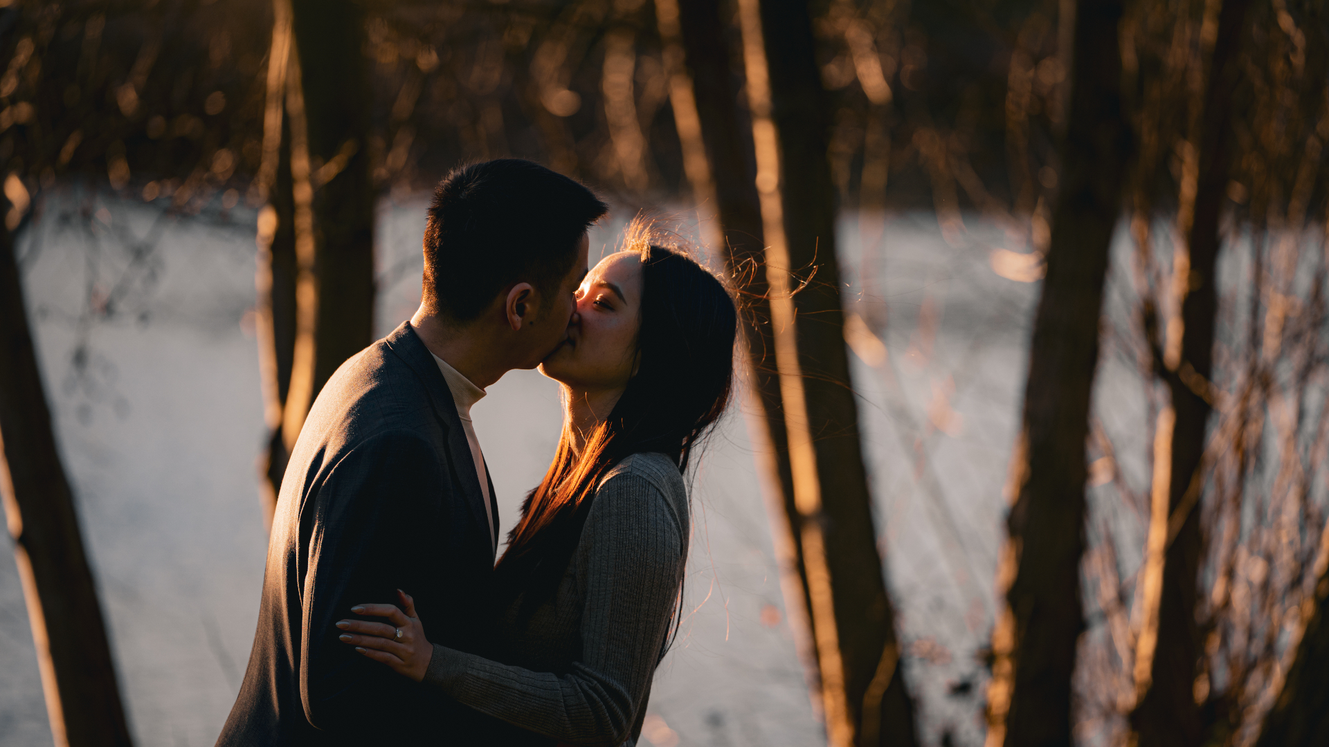 Engaged couple kissing through silhouetted winter trees at sunset