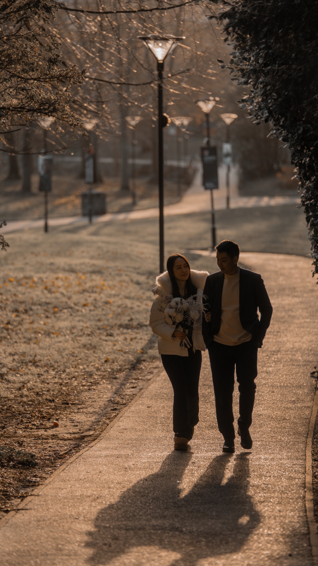 Newly engaged couple walking down a sunlit park path with bouquet