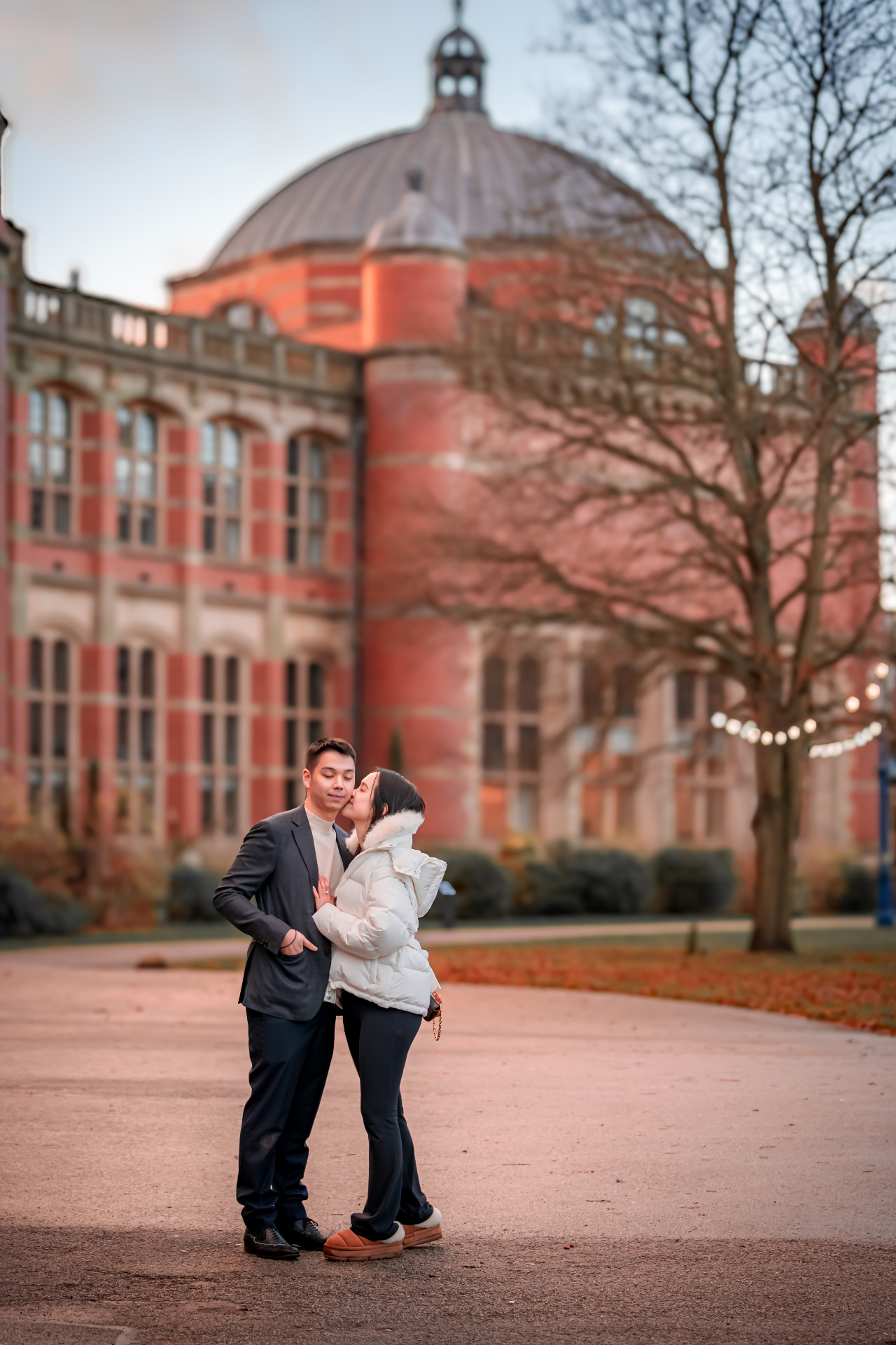 Bride-to-be kissing fiancé's cheek in front of red brick dome building